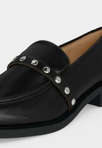 Mocassin en cuir noir avec un petit talon et une bride ornée de clous ronds argentés sur le dessus.