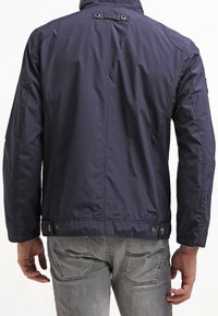 Marineblauwe, lichte jas met een staande kraag en knoopaccenten, uitgevoerd in een licht getextureerde stof en met een ontspannen pasvorm. Grijze jeans zichtbaar.