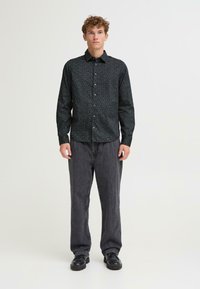 Camicia nera a maniche lunghe con piccolo motivo bianco, chiusura con bottoni e colletto, abbinata a pantaloni grigi e scarpe nere.