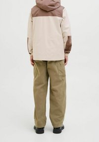 Giacca beige chiaro con dettagli marroni, cappuccio e polsini elastici; abbinata a pantaloni verdi oliva larghi e scarpe nere. Sfondo semplice.