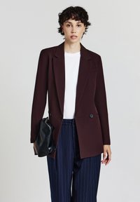 Giovane con capelli ricci e corti, indossa un blazer marrone scuro, una camicia bianca e pantaloni blu a righe. Sta tenendo una pochette nera.