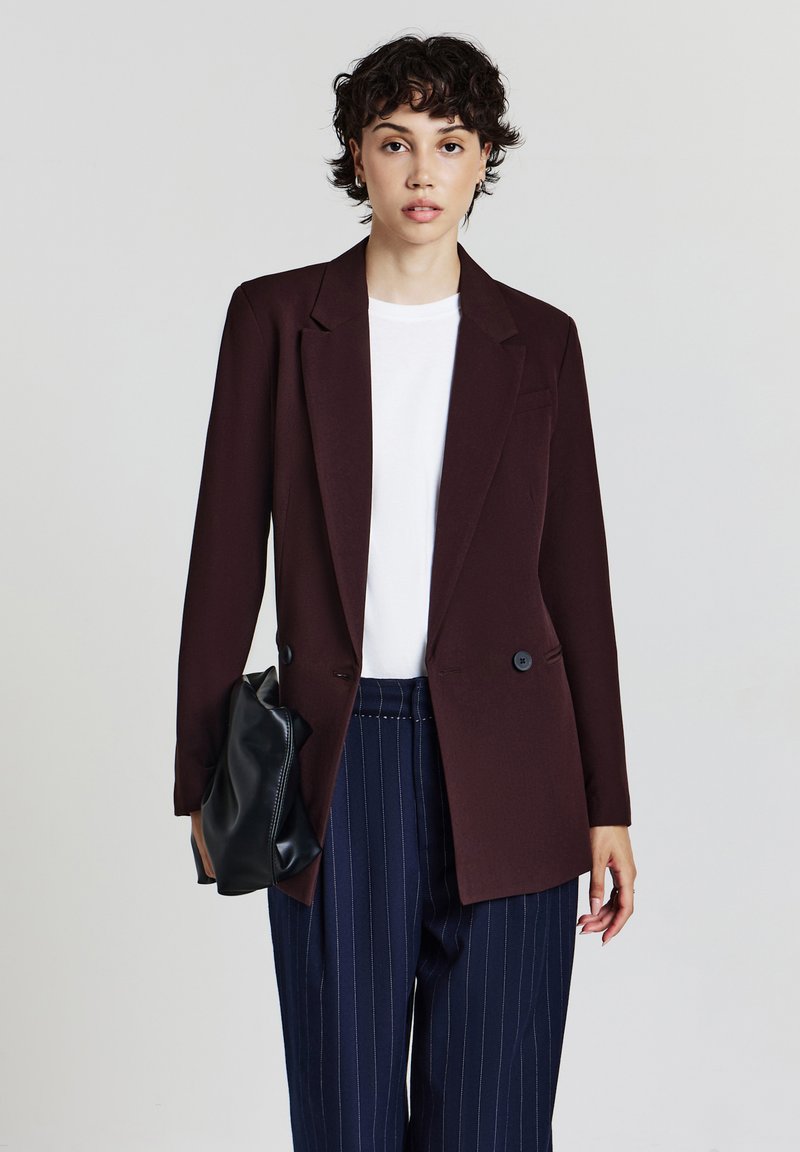 Giovane con capelli ricci e corti, indossa un blazer marrone scuro, una camicia bianca e pantaloni blu a righe. Sta tenendo una pochette nera.