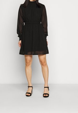 Vrouw die een zwart knielang jurkje draagt met doorschijnende lange mouwen en zwarte hoge hakken met enkelbandje, staand tegen een effen achtergrond.