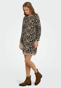 Femme portant une robe à manches longues beige et noire à imprimé léopard et des bottes brunes à mi-mollet, debout sur un fond blanc.