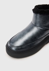 Fekete bőr bokacsizma vastag talppal és a tetején látható fekete szőr béléssel, közelről bemutatva fehér háttér előtt.