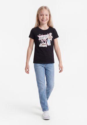 Ragazza sorridente che cammina in avanti indossando una t-shirt nera con grafica, jeans azzurri chiari e scarpe bianche su uno sfondo bianco uniforme.