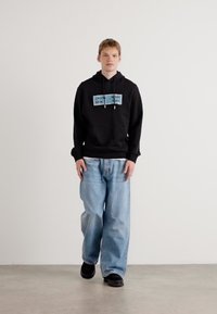 Zwarte hoodie met twee blauwe merknamen op de borst, gecombineerd met losvallende lichtblauwe jeans en zwarte schoenen. Effen achtergrond.