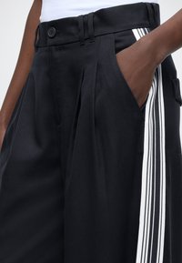 Črne hlače z naborki in zapenjanjem na gumbe na pasu, z bočnima žepoma in kontrastnim belo navpično črto ob strani.