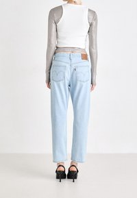 Persoon in een metalen langemouwen top onder een witte mouwloze crop top, lichtblauwe high-waisted jeans en zwarte hoge hakken, staat.