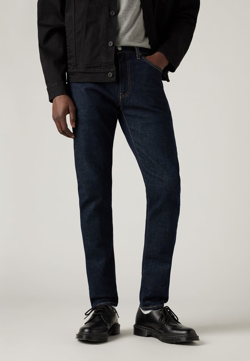 Hombre vestido con vaqueros oscuros, zapatos de cuero negros, camisa gris y chaqueta negra, con una mano en el bolsillo, frente a un fondo neutro.