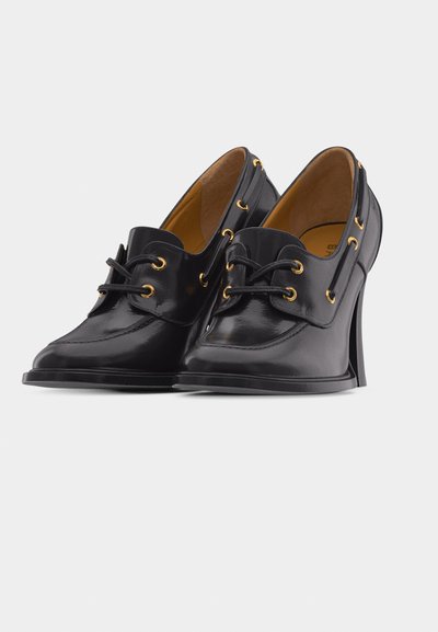 Μαύρα δερμάτινα ψηλοτάκουνα παπούτσια με κορδόνια μπροστά, χρυσά εξωτερικά κουμπώματα και καμπυλωτό σχέδιο τακουνιού σε απλό λευκό φόντο.
