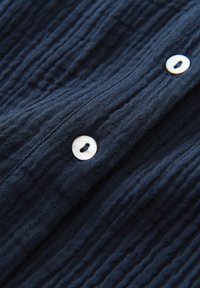 Tessuto texture blu navy con increspature visibili, caratterizzato da un pulsante rotondo e iridescente. Focus sulla trama del tessuto e sui dettagli del pulsante.
