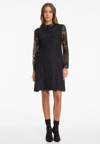 Black lace dress with long transparent sleeves, a scalloped hem, and a rounded neckline. The material is intricate with floral patterns.