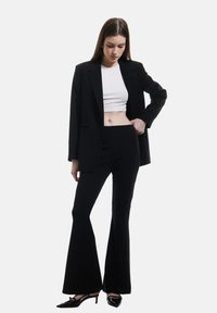 Blazer nero su misura sopra un crop top bianco, abbinato a pantaloni neri a campana e tacchi alti neri. Tessuto morbido, vestibilità strutturata, stile minimalista.