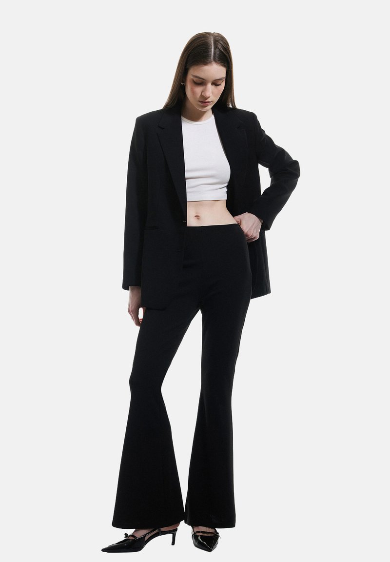 Blazer nero su misura sopra un crop top bianco, abbinato a pantaloni neri a campana e tacchi alti neri. Tessuto morbido, vestibilità strutturata, stile minimalista.
