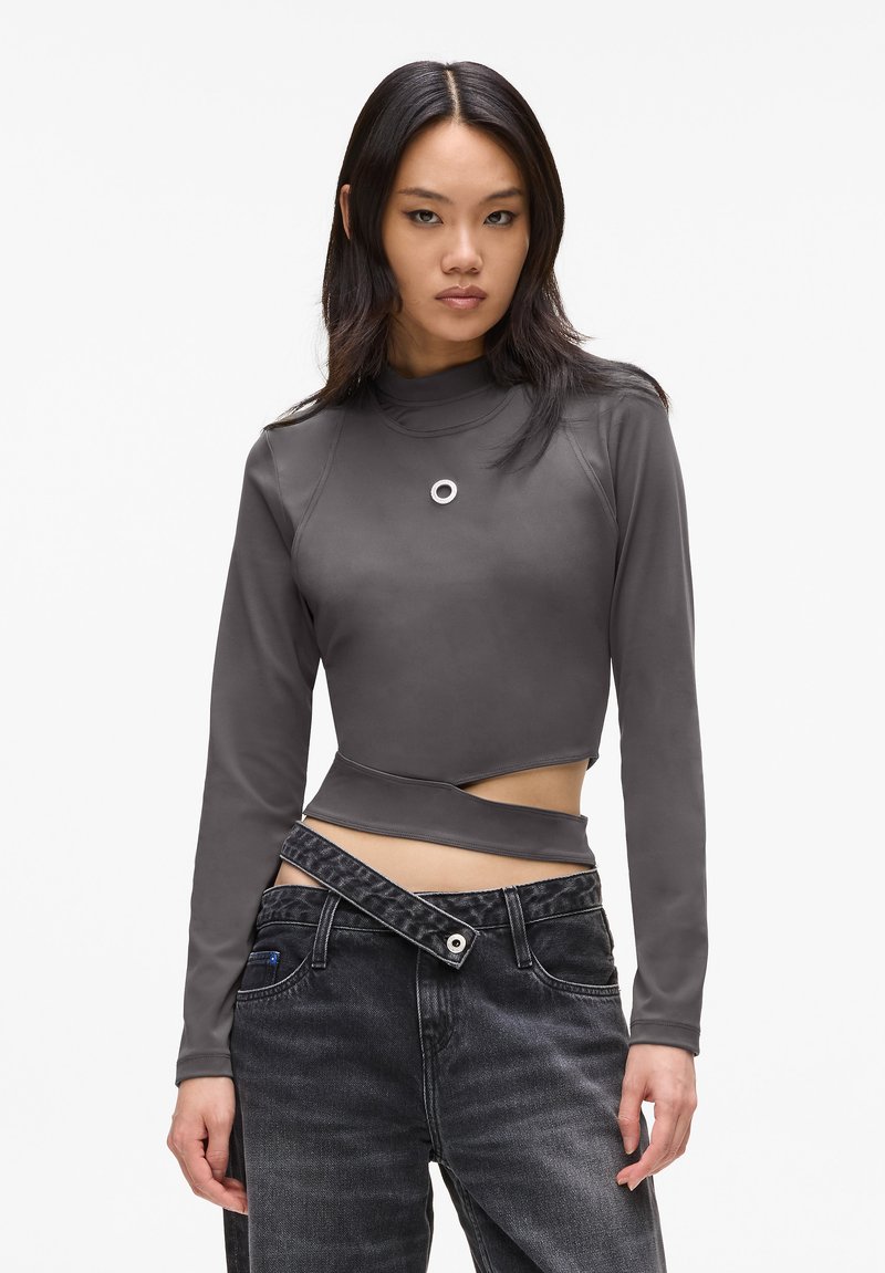 Maglione grigio a maniche lunghe con collo alto, caratterizzato da un design asimmetrico e un accento circolare. Abbinato a jeans scuri a vita alta.