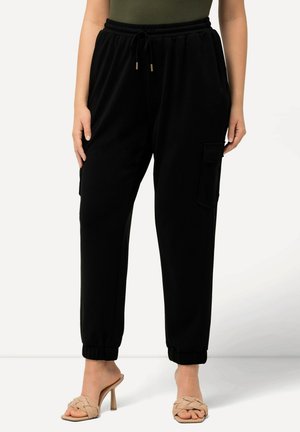 Pantaloni cargo neri con vita elasticizzata e coulisse, tasche laterali e polsini elasticizzati, abbinati a sandali con tacco color sabbia. Texture liscia.
