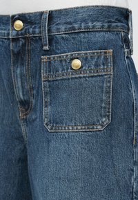 Tumši zili džinsu bikses ar zelta metāla pogām, kontrastējošu diegu un mazu taisnstūrveida priekšējo kabatu.