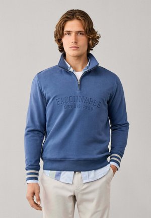 Jeune homme aux cheveux ondulés porte un pull bleu à fermeture éclair quart avec des poignets rayés sur une chemise rayée et un pantalon clair, debout droit.
