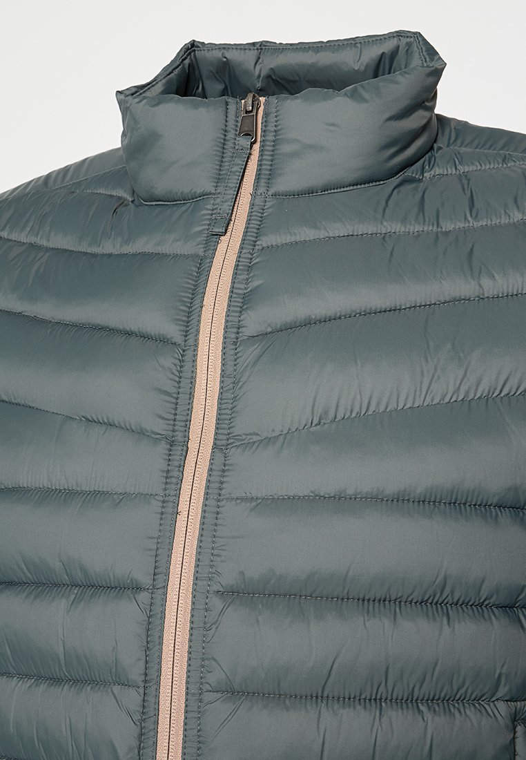 Veste matelassée en teinte bleu canard, dotée d'un col haut et d'une fermeture éclair beige contrastante. Le tissu semble lisse et léger.