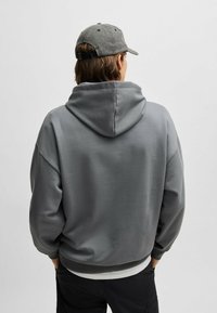 Grijze pullover hoodie met een grote capuchon, ribgebreide manchetten en een ontspannen pasvorm, vanachter getoond, gecombineerd met een grijze pet.