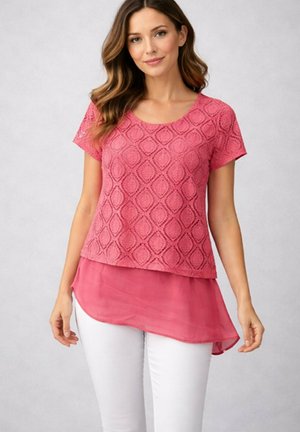 Femme portant un haut rose à manches courtes à motifs avec un ourlet asymétrique transparent, associé à un pantalon blanc, debout devant un fond uni.