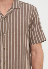 Καφέ πουκάμισο με κοντά μανίκια και κάθετες λευκές ρίγες, φορεμένο από ένα άτομο μπροστά σε απλό φόντο.