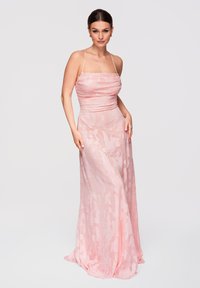 Vrouw draagt een lang, roze satijnen jurk met dunne bandjes, staand met één hand op haar heup tegen een effen witte achtergrond.