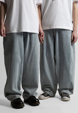 Dvaja ľudia oblečení v bielych tričkách a nadrozmerných svetlomodrých džínsoch, jeden s čiernymi topánkami a druhý s čierno-bielymi teniskami.