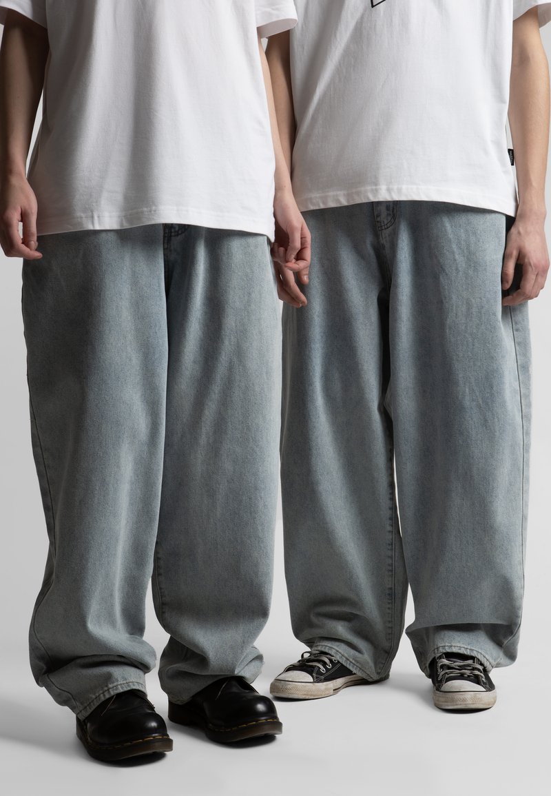 Δύο άτομα φοράνε λευκές μπλούζες και φαρδιά ανοιχτόμπλε τζιν, το ένα με μαύρες μπότες και το άλλο με μαύρα και άσπρα αθλητικά παπούτσια.