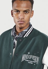 Chaqueta varsity verde con cuello de canalé a rayas negras y blancas, que presenta el texto bordado "MIND OF VISION" y una superficie de tejido texturizado.