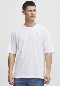 Hvid bomulds t-shirt med rund hals og korte ærmer. Har navy "A.R.C." tekst på venstre side. Bæres med blå jeans.