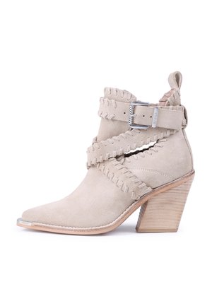 Beige suede ankle boot with pointed toe, layered straps, decorative stitching, and a stacked wooden heel. Metallic buckle accent.