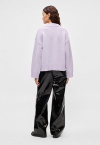 Λιλά πλεκτό πουλόβερ με ριμπ τελείωμα, φορεμένο με μαύρο, γυαλιστερό, φαρδύ παντελόνι. Το πουλόβερ έχει χαλαρή εφαρμογή και κατεβασμένους ώμους.