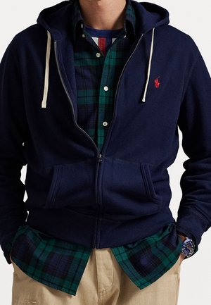 Hombre con sudadera con cremallera azul marino sobre camisa de cuadros verde y azul marino, pantalones beige y reloj de pulsera azul, con las manos en los bolsillos.