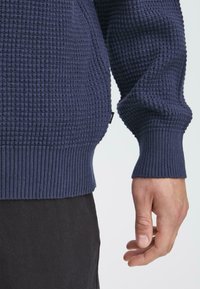 Pull en maille texturée marine avec un motif gaufré ; poignets et ourlet côtelés, affichant un petit label sur le côté. Main posée sur l'ourlet.