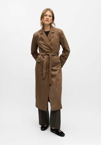 Mujer de pie con las manos detrás de la espalda, vistiendo un abrigo largo marrón con cinturón, pantalones oscuros y zapatos negros, contra un fondo blanco.