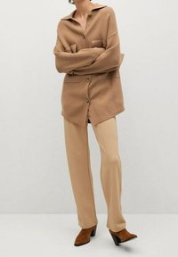 Camicia beige oversize con bottoni e due tasche frontali, abbinata a pantaloni beige chiaro sartoriali e stivaletti con tacco marroni.