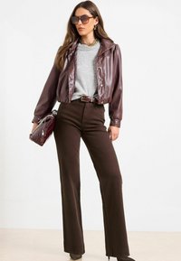 Hnědá kožená bunda na cropped střih, šedý svetr, hnědé zvonové kalhoty a oversized sluneční brýle. Zlatý náhrdelník a kabelka s řetízkovým popruhem.