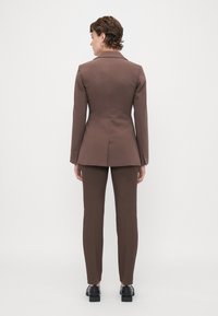 Hnědý zpevněný blazer s vykrojenými klopami a přiléhavými kalhotami. Hladká tkanina, přiléhavá struktura a jemné švy. Černé boty.