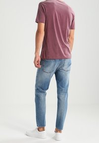 Mężczyzna w wyblakłych niebieskich dżinsach, białych sneakersach i krótkim, bordowym t-shircie, stojący tyłem na białym tle.