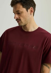 Bordowa koszulka z okrągłym dekoltem, wykonana z miękkiego materiału, z wypukłym logo na przodzie. Krótkie rękawy i luźny krój.