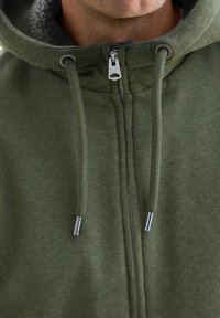 Felpa verde con zip, dotata di un morbido rivestimento interno, hardware argentato e cordini regolabili sul cappuccio, con una finitura in tessuto texturizzato.
