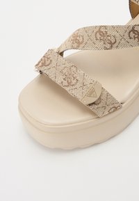 Beige Plattform-Sandale mit gemusterten Textilelementen und einer glatten, strukturierten Sohle. Metallene Logoakzente verleihen dem Design zusätzlichen Ausdruck.