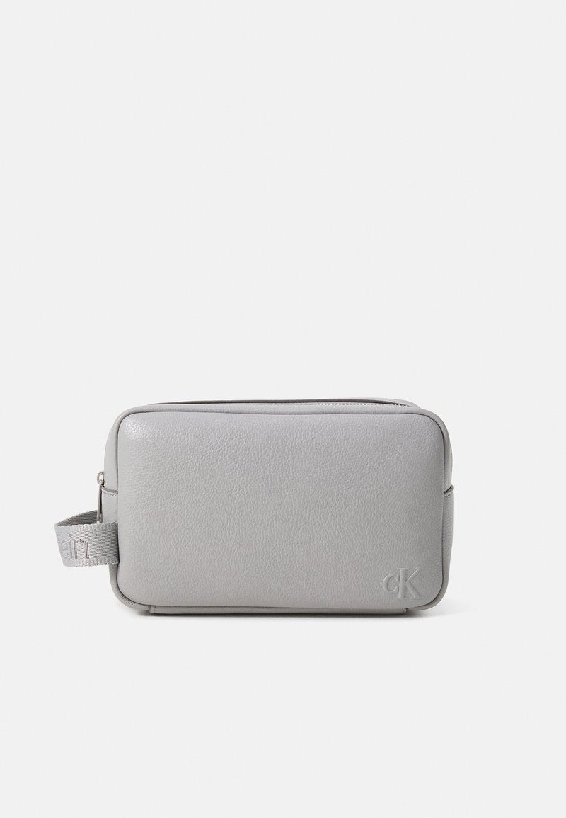 Trousse de maquillage en cuir gris au format rectangulaire, avec une fermeture zippée sur le dessus, une poignée sur le côté et un détail de logo embossé à l'avant.