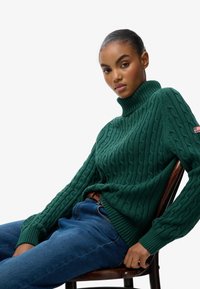 Zielony sweter z wysokim kołnierzem w sploty, z prążkowanym wykończeniem, zestawiony z niebieskimi dżinsami. Sweter ma teksturowany wzór oraz mały akcent z logo na rękawie.