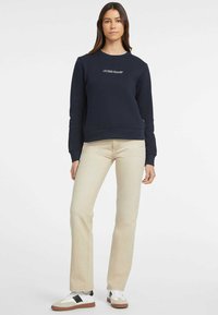 Felpa navy con logo bianco "Guess Jeans", scollatura rotonda, polsini e orlo a costine, abbinata a jeans beige chiaro a gamba dritta e sneakers.
