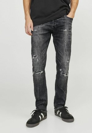 Homme portant un jean noir déchiré avec des déchirures et des éclaboussures de peinture, des baskets noires avec des rayures blanches et une chemise noire avec une main dans la poche.