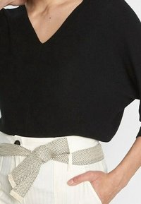 Czarna dzianinowa bluzka z dekoltem w serek i luźnym krojem, noszona z jasnymi, wysokimi spodniami z paskiem w wzór rybiej ości.