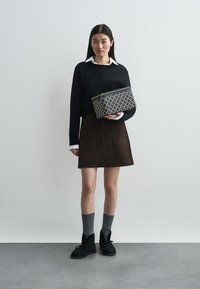 Kvinna i svart tröja, vit skjorta med krage, brun kjol, grå strumpor, svarta stövlar, håller en rektangulär väska med svart och vitt mönster.